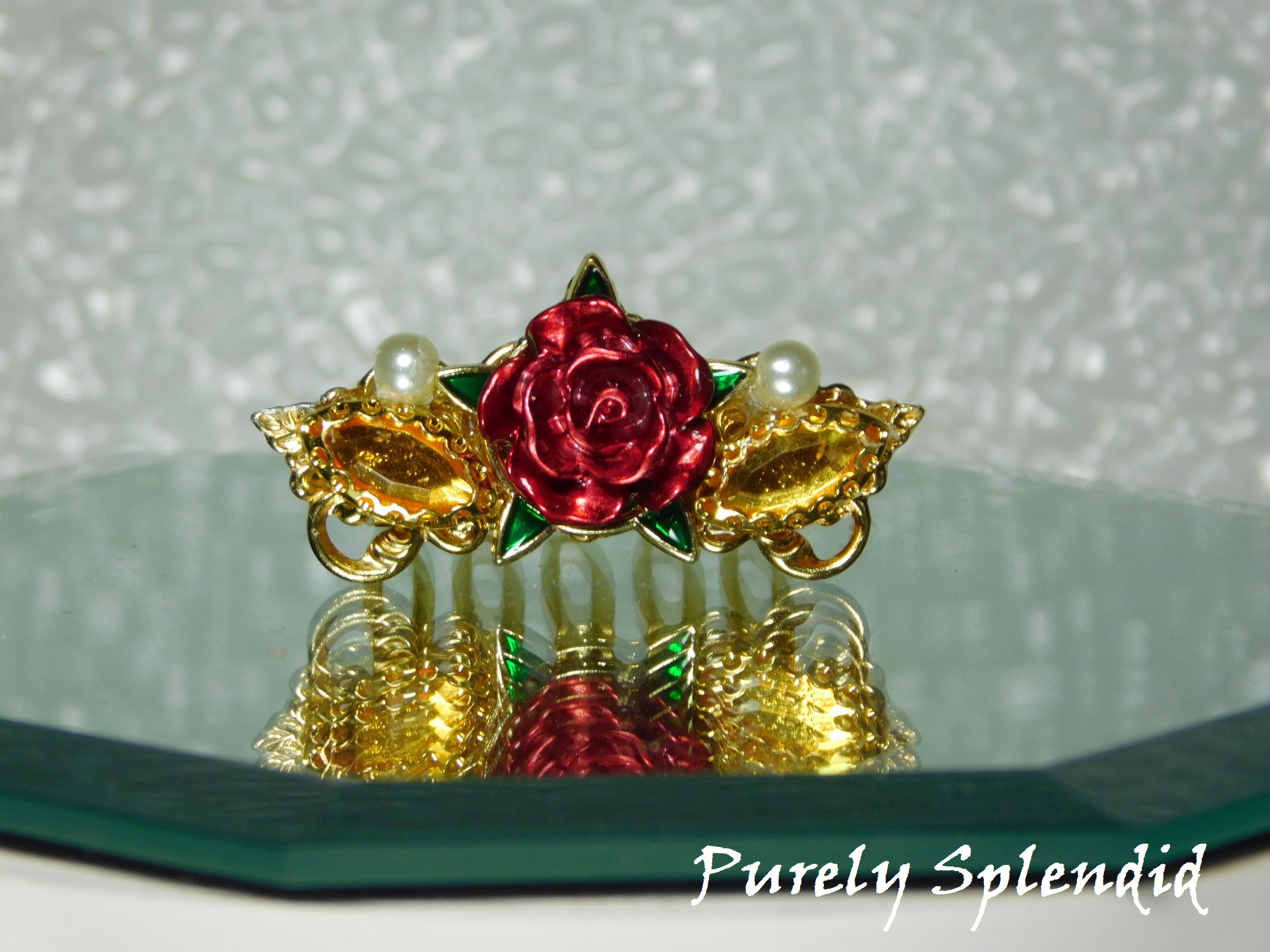 large center  rose with five green leaves and a gold colored horse eye rhinestone and small white pearl on either side of the rose 