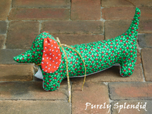 Load image into Gallery viewer, dachshund made of green fabric with green holly leaves and red heart berries, ears of red fabric with white hearts and green leaves and a white under body wearing a thin gold metallic bow around the neck and a silver heart charm hanging from it
