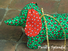 Load image into Gallery viewer, dachshund made of green fabric with green holly leaves and red heart berries, ears of red fabric with white hearts and green leaves and a white under body wearing a thin gold metallic bow around the neck and a silver heart charm hanging from it
