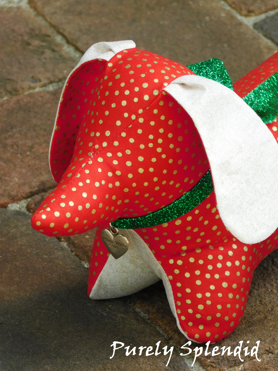 dachshund made of red fabric with gold dots, light colored ears and under body with a sparkling green bow tied around the neck and a silver heart dangling from it