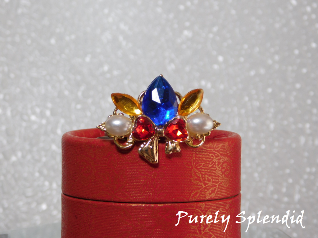 Snow White Crown for dolls with a large blue center stone, sparkling red bow with white pearls on either side of it sitting on a red gift box