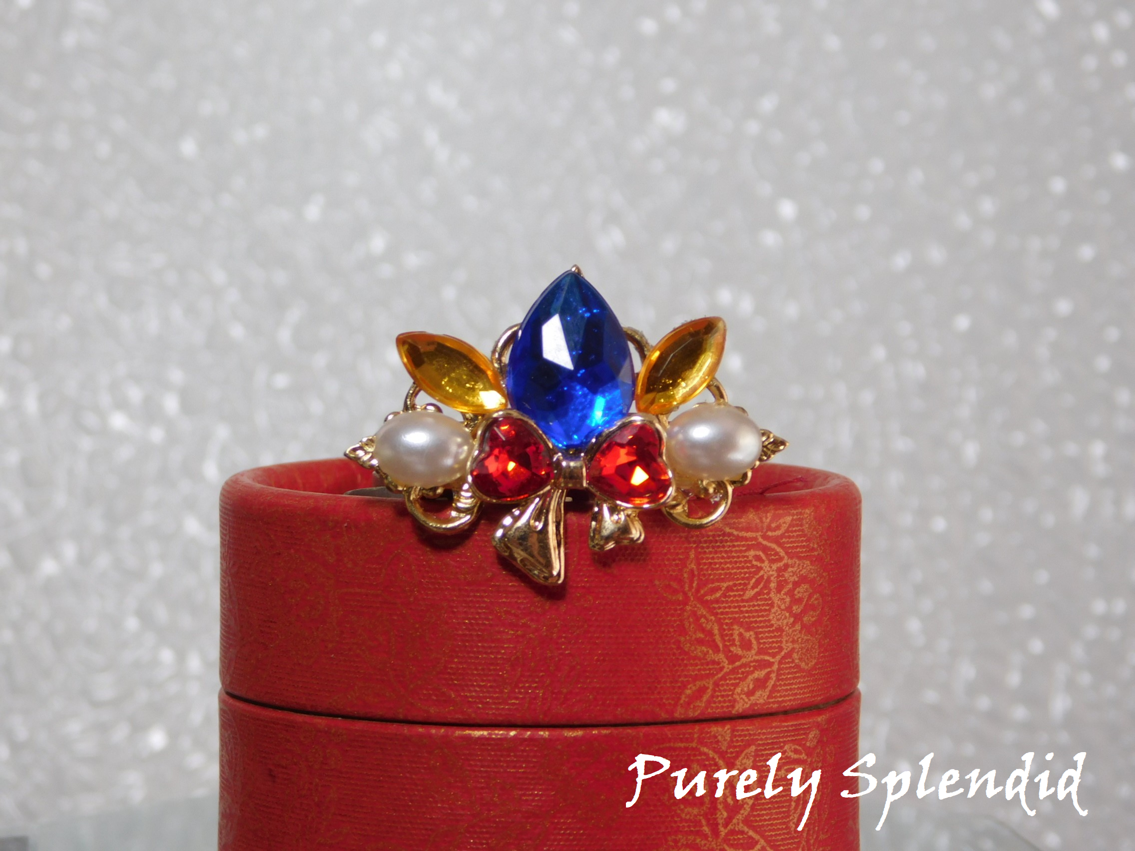 Snow White Crown for dolls with a large blue center stone, sparkling red bow with white pearls on either side of it sitting on a red gift box