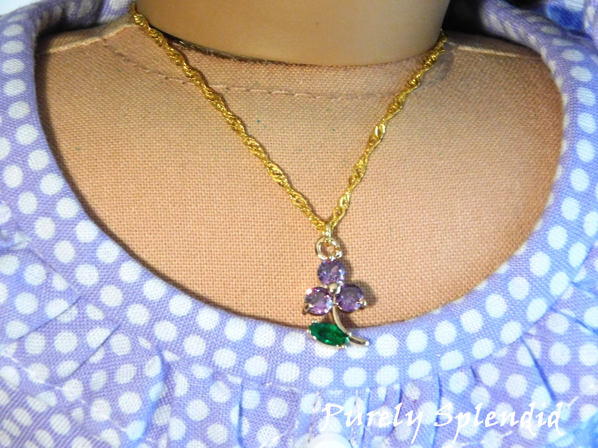 18 inch doll shown wearing a Sparkling Purple Flower Necklace. Flower charm has 3 sparkling purple petals and a sparkling green leaf hanging from a pretty twisted gold chain.