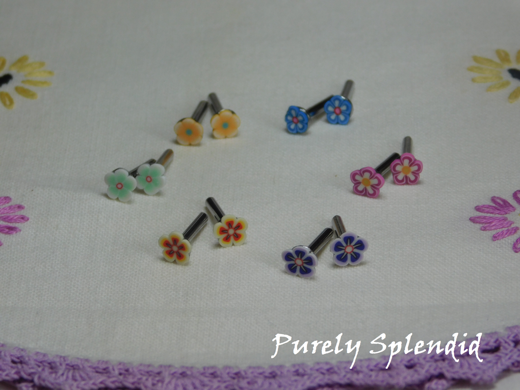 Six pairs of colorful Flower Studs shown on a white floral background. Mint green flowers with pink centers, light orange flowers with light blue centers, blue flowers with dark orange centers, pink flowers with yellow centers, purple flowers with yellow centers and yellow and orange flowers with yellow centers 