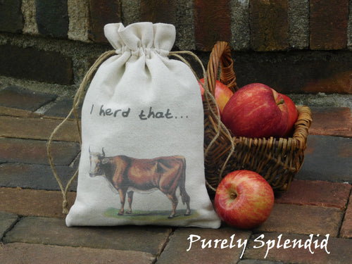 fabric gift bag with a vintage image of a brown cow and the words I herd that. Bag is cinched closed and sitting in front of a basket of apples on a brick step