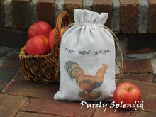 Load image into Gallery viewer, Fabric gift bag with an image of a vintage rooster and the words rise and shine. shown in front of a basket of apples on a brick step. Bag is cinched closed
