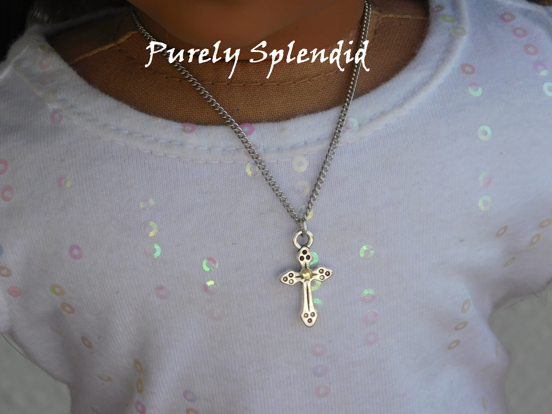 Decorative Silver Cross with a clear sparkling center crystal hanging from a silver chain around an 18 inch dolls neck