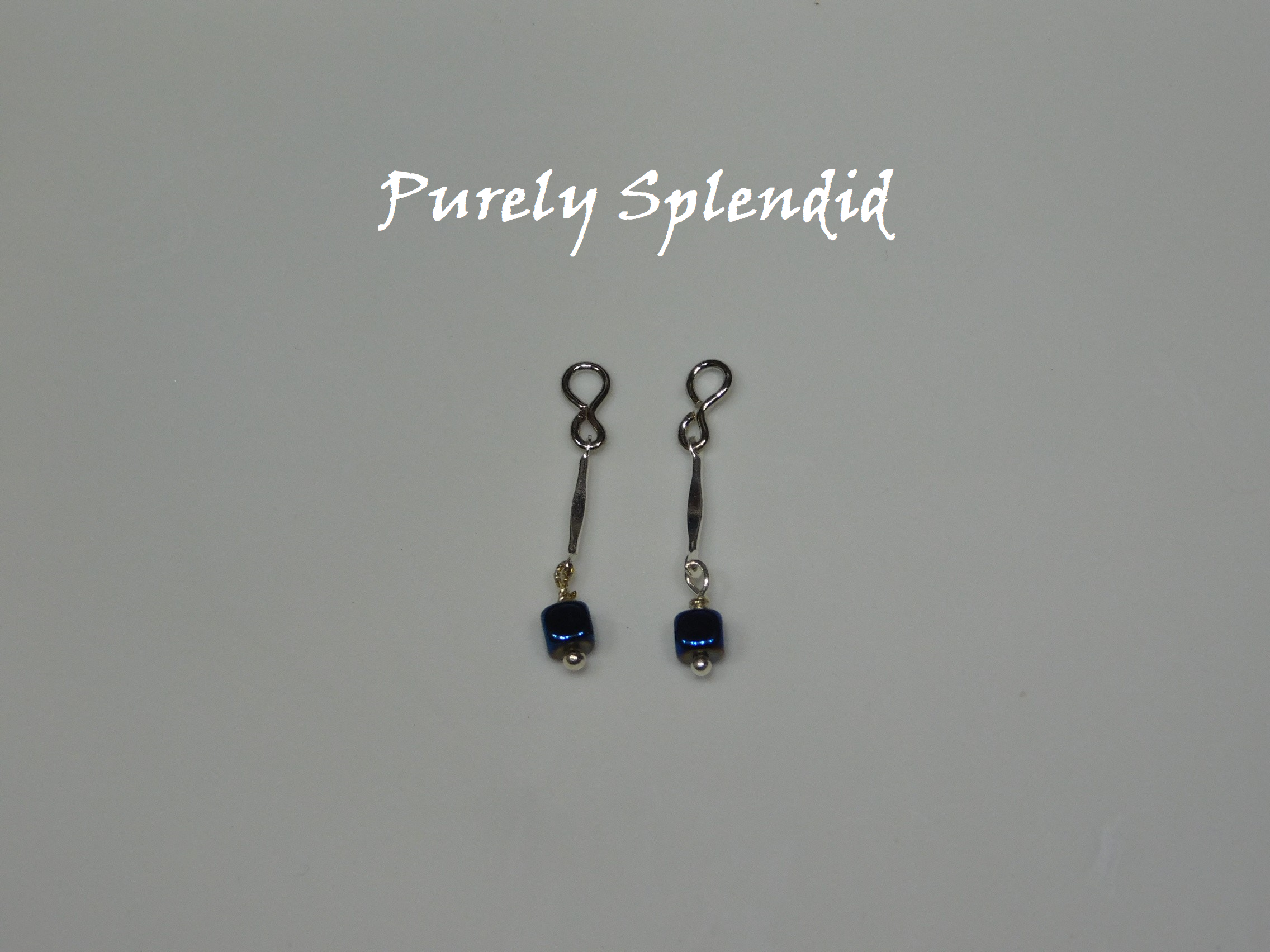 Long thin silver bead with a dark blue square bead dangling below. Shown on a white background