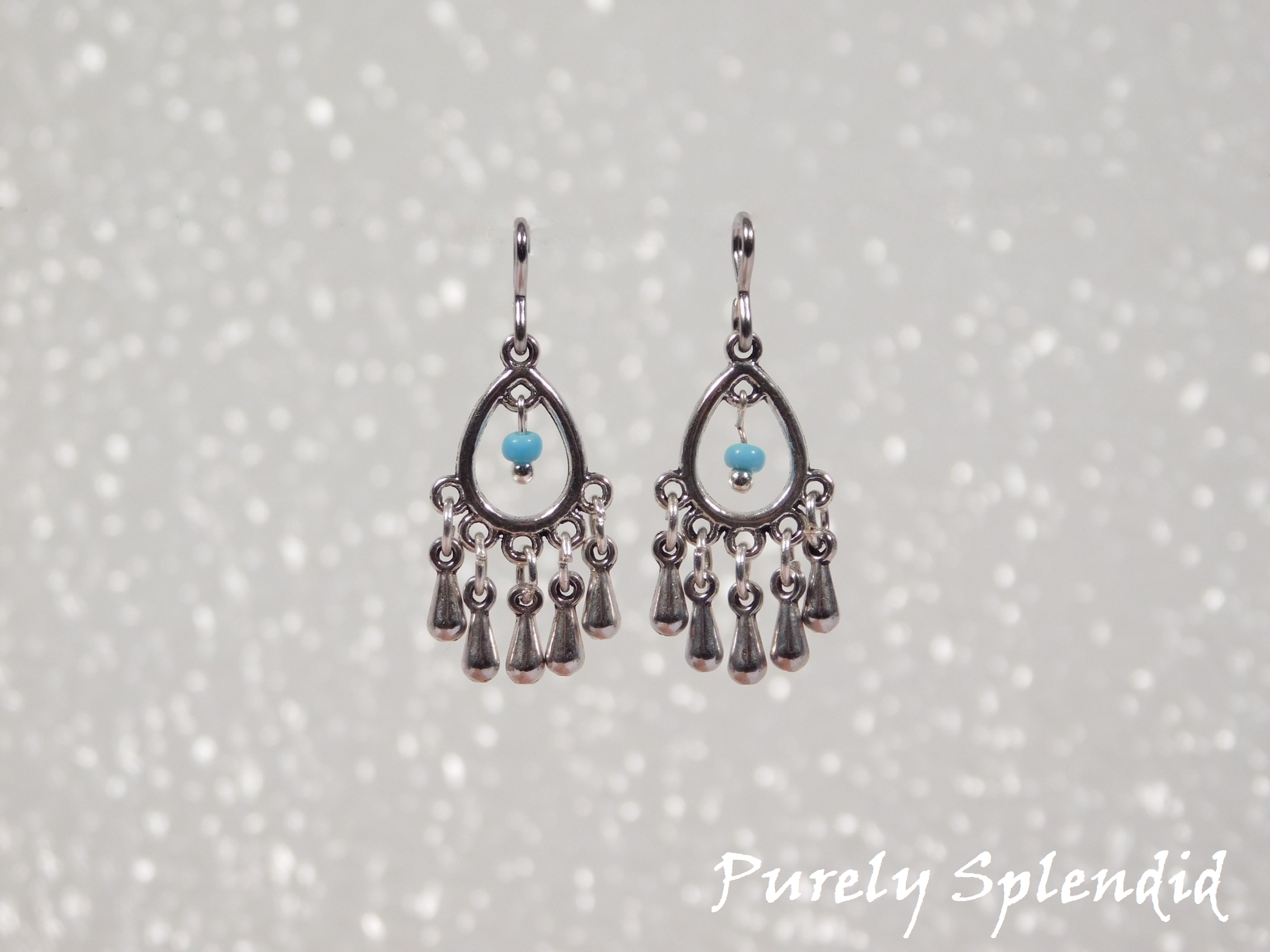 Silver colored open teardrop shaped earrings with five silver colored drops hanging below and one blue colored bead hanging in the center of the teardrop.