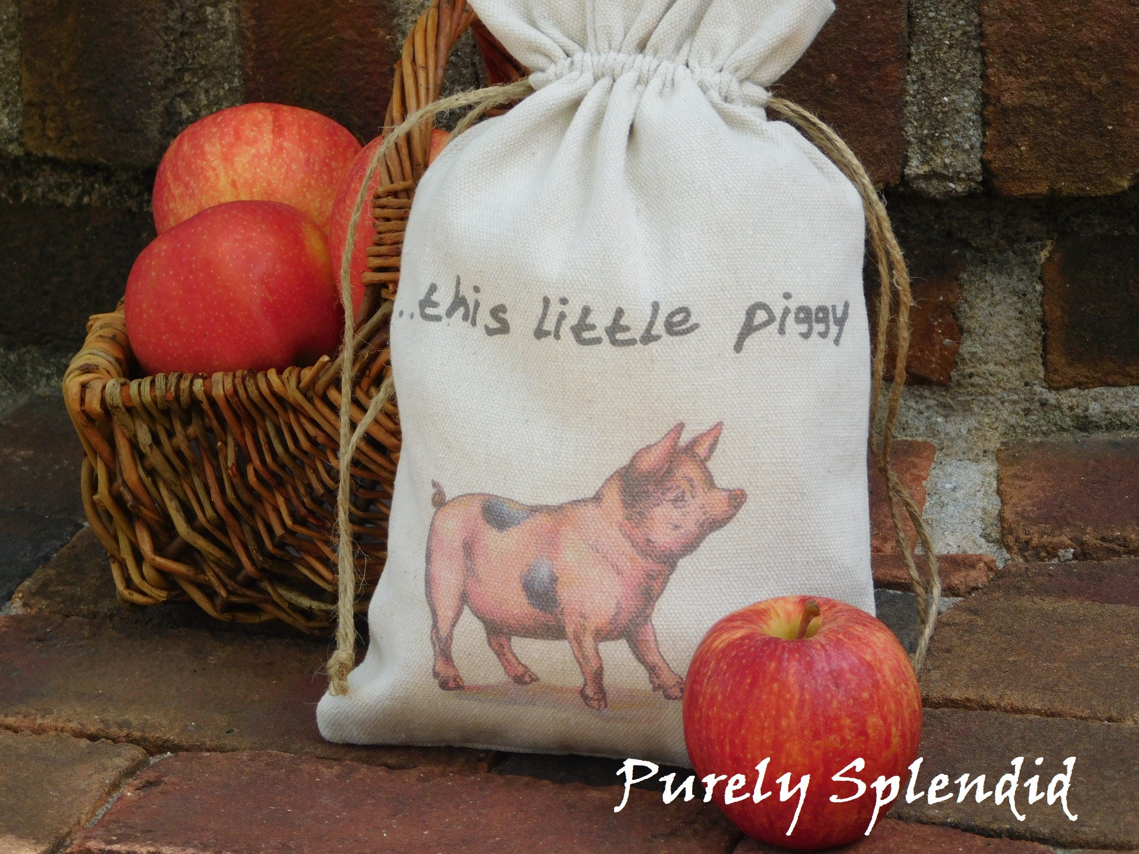 close up picture of a vintage style pink pig fabric bag with the words ...this little piggy. Bag is cinched up sitting in front of a basket of apples
