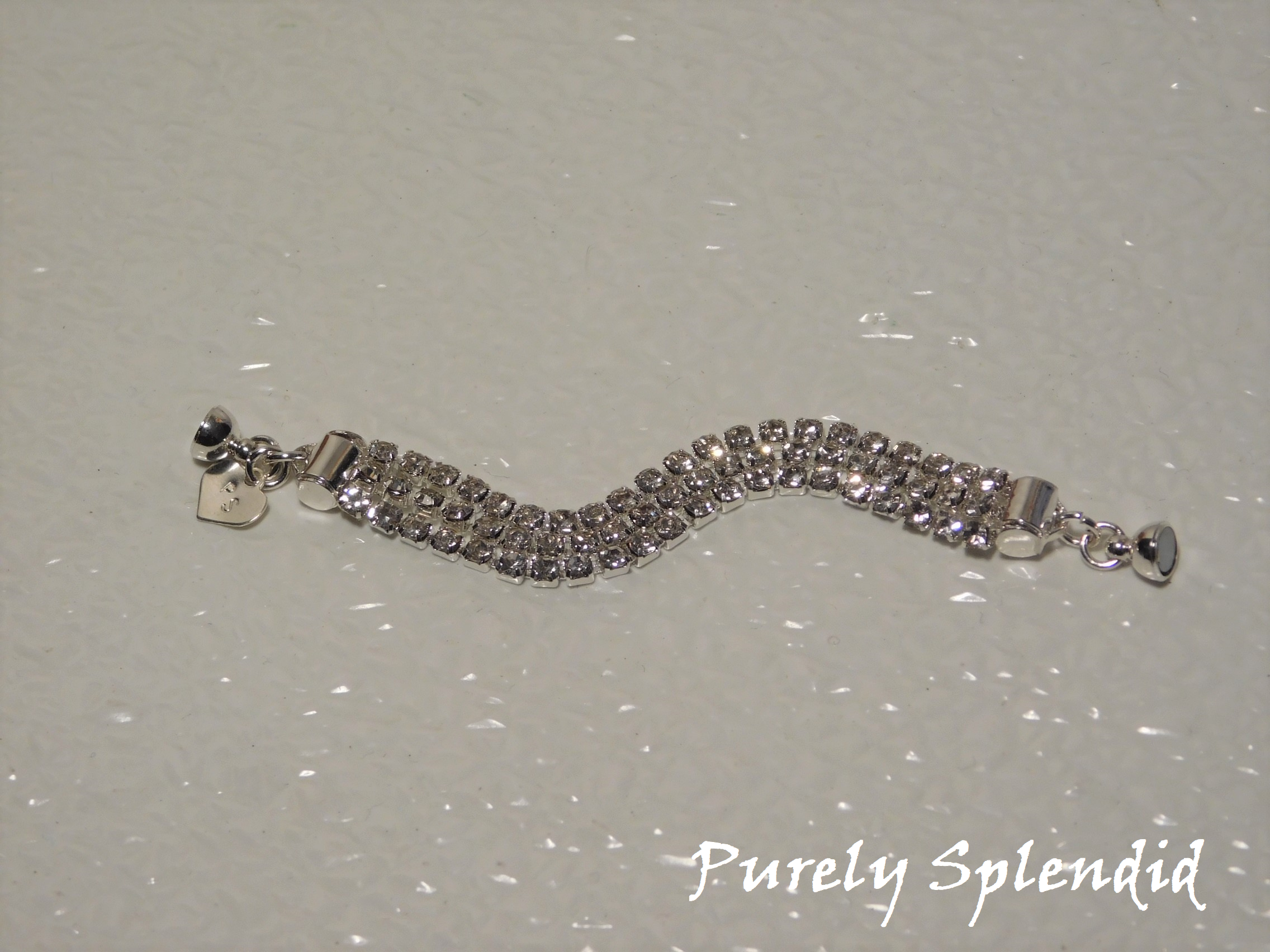 Three rows of silver colored rhinestones make up this Triple Silver Rhinestone Bracelet. Closes with a magnetic clasp. Shown laying on a white background