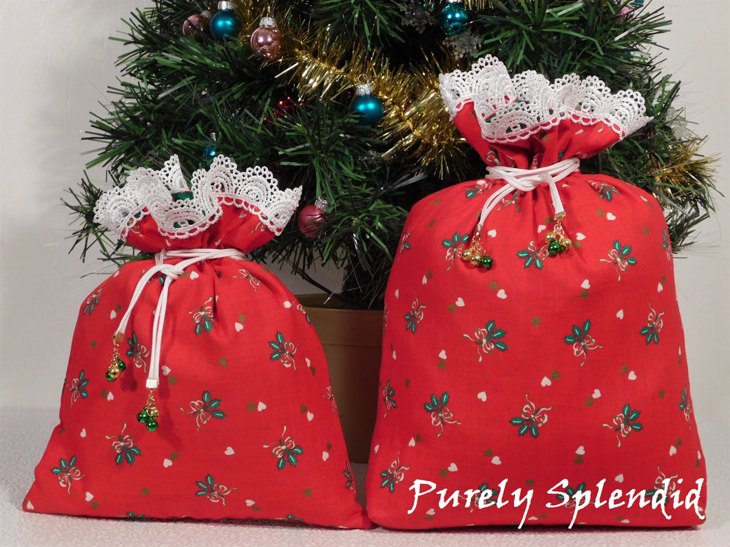 two red Christmas themed fabric gift bags shown with white lace edging the tops, each tied with white drawstrings that have tiny jingle bells at the ends.